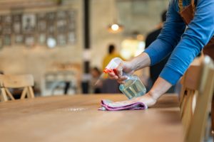 Mulher limpando uma mesa de madeira. Em sua mão direita, há um borrifador e na esquerda, uma flanela.